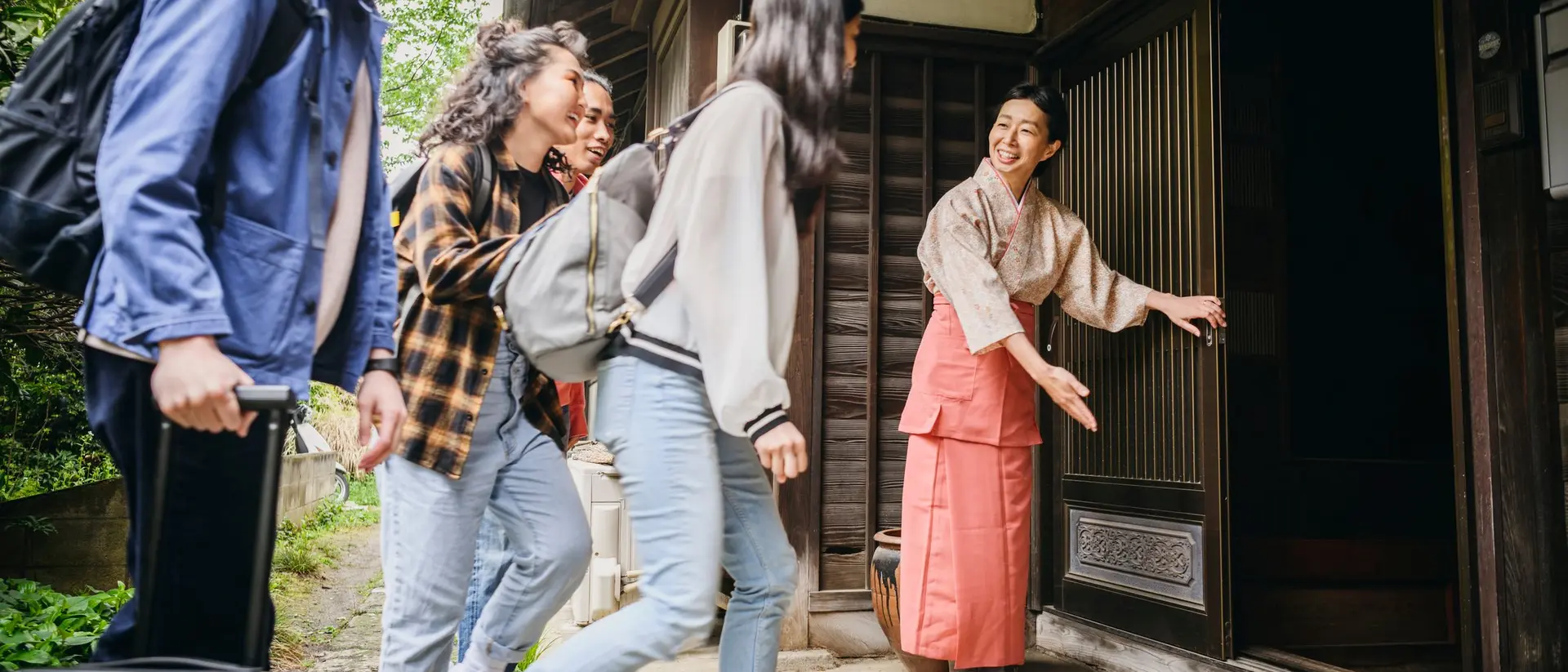 Four young adults approach a traditional home, greeted by a smiling woman in a kimono at the entrance.