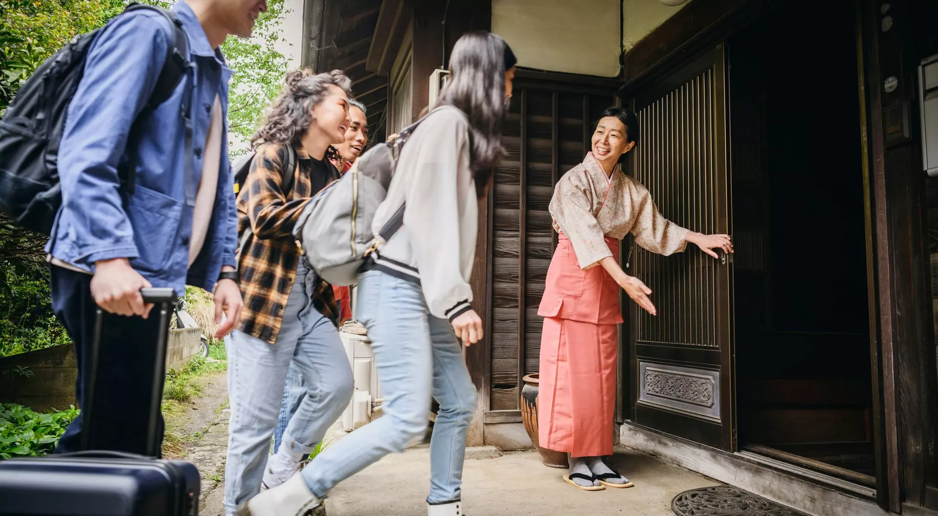 Four young adults approach a traditional home, greeted by a smiling woman in a kimono at the entrance.