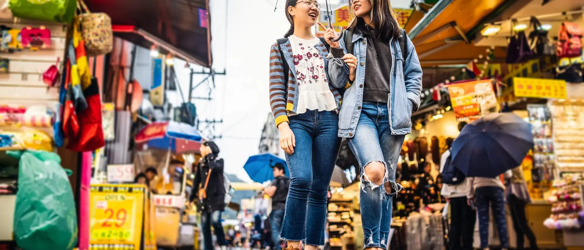 Two friends walk together under an umbrella in a vibrant market, surrounded by colorful stalls and wet pavement.