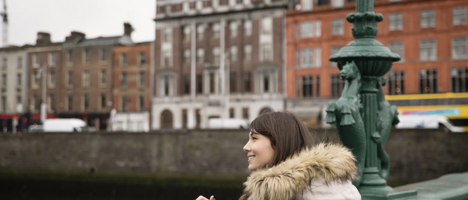 A woman in a cozy coat stands by a river, smiling and looking at buildings in the background on a cloudy day.