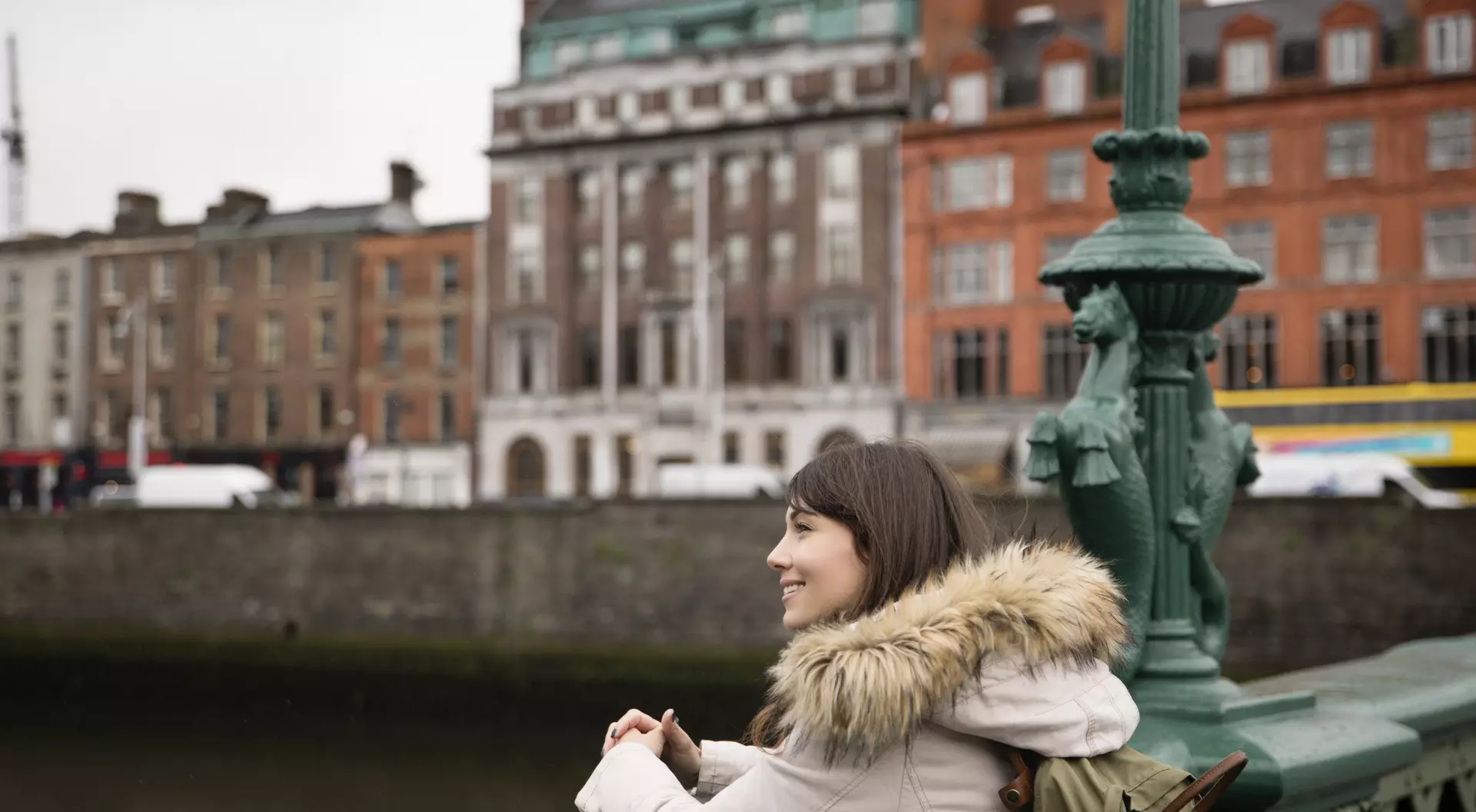 A woman in a cozy coat stands by a river, smiling and looking at buildings in the background on a cloudy day.