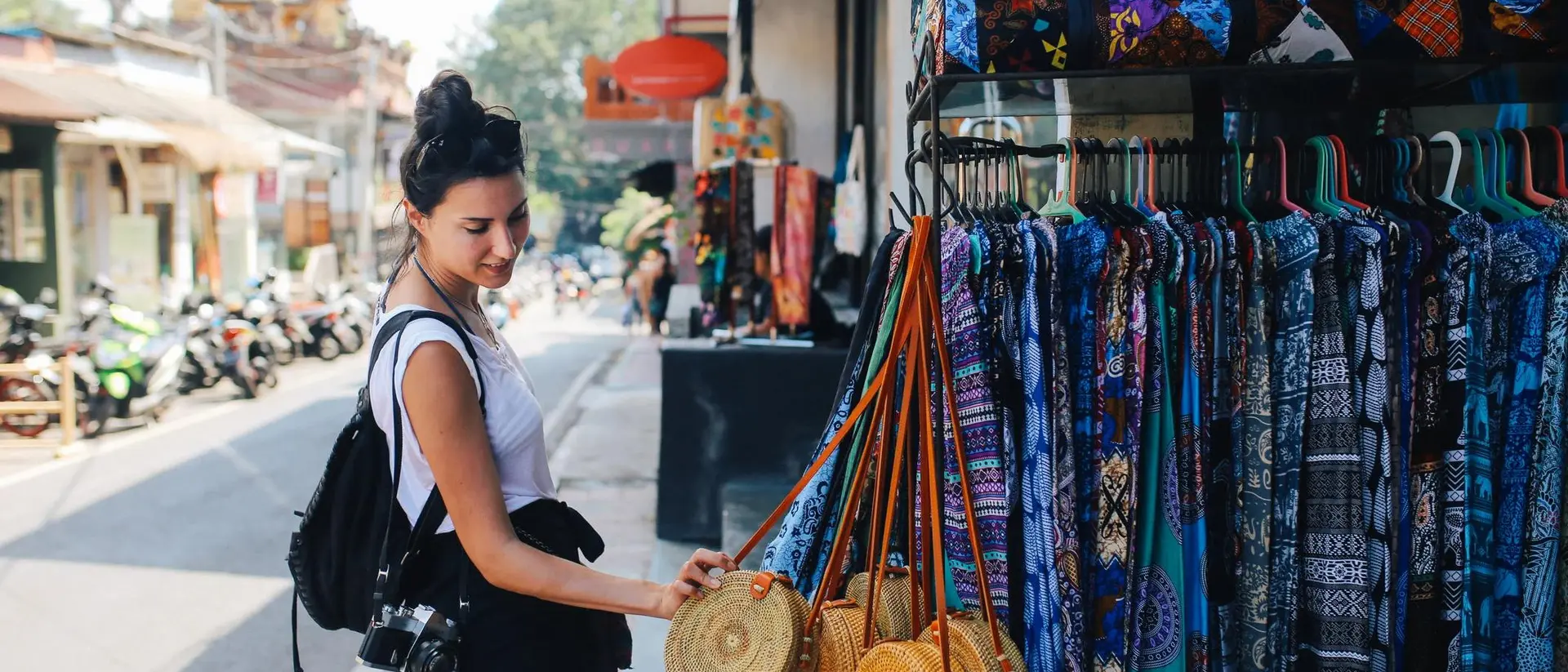 A woman examines woven bags in a market, with colorful fabrics hanging nearby and a street scene in the background.