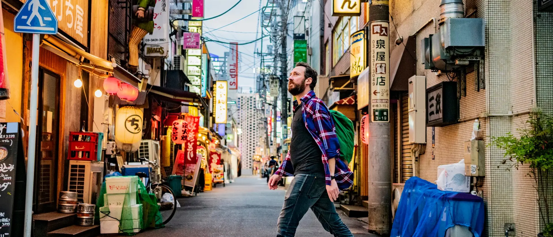 A man in a plaid shirt and red sneakers walks across a crosswalk in a vibrant city street filled with neon signs.