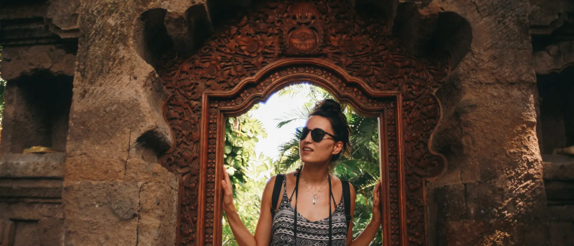 A person in sunglasses stands in a carved stone doorway, surrounded by greenery. A camera hangs from their neck.