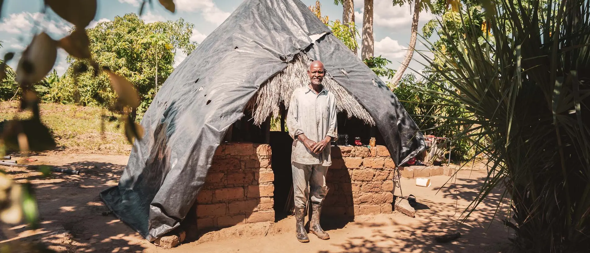 Man standing in front of a small mud brick house with a thatched roof in a natural outdoor setting.
