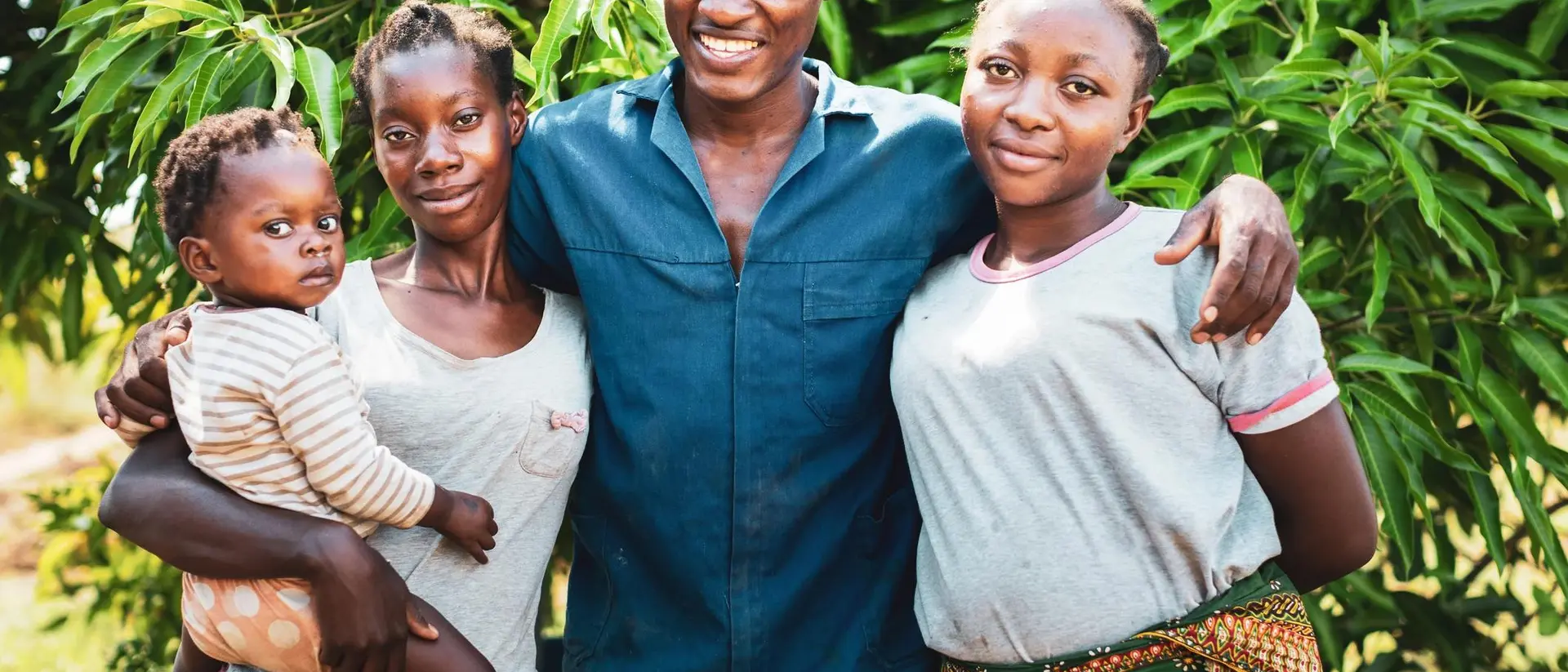 A smiling man stands with three women, one holding a baby, in front of lush green foliage.