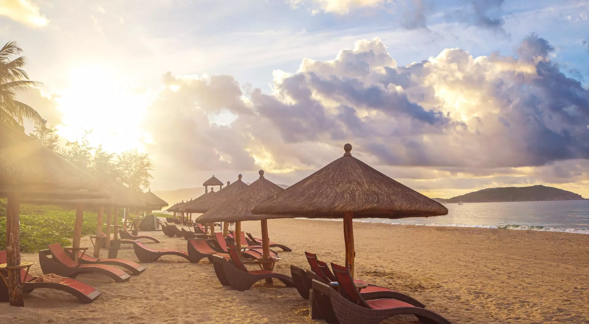 Beach scene with lounge chairs under straw umbrellas, illuminated by a warm sunset and scattered clouds.