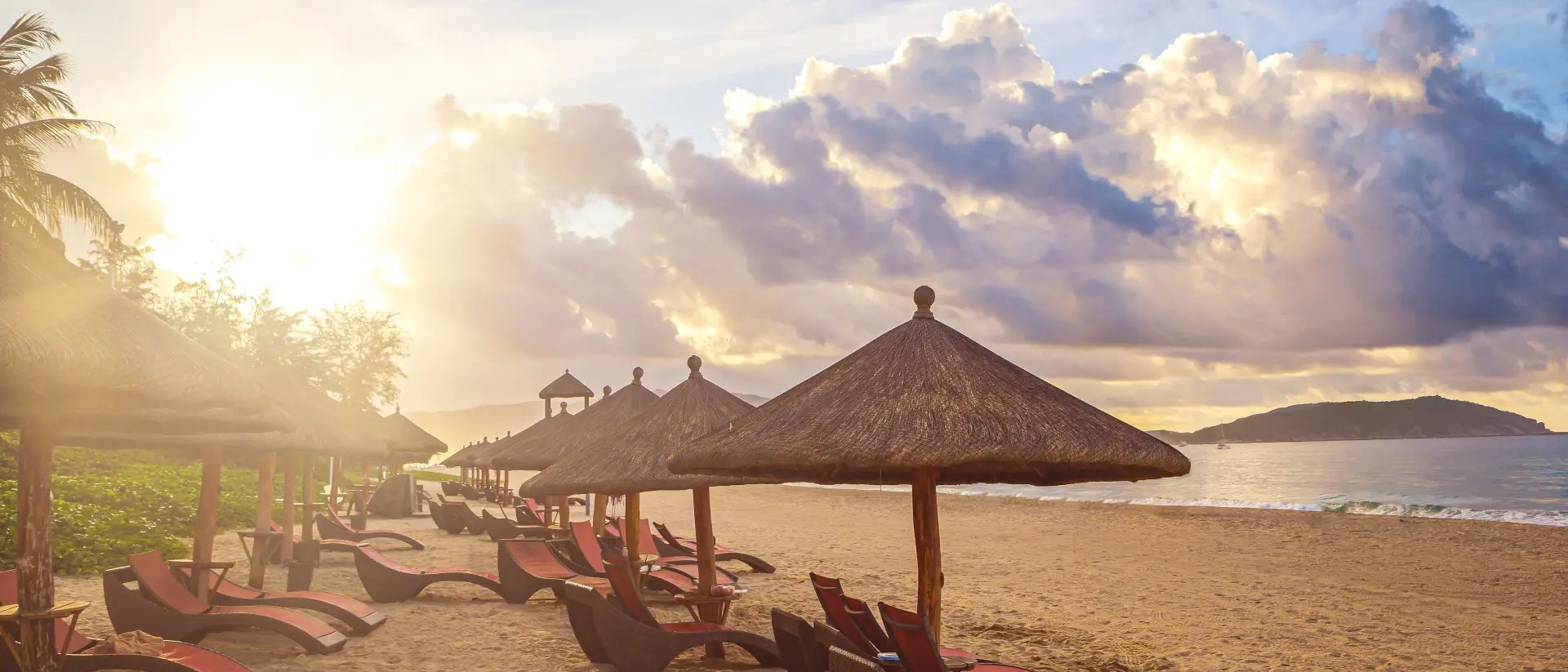 Beach scene with lounge chairs under straw umbrellas, illuminated by a warm sunset and scattered clouds.
