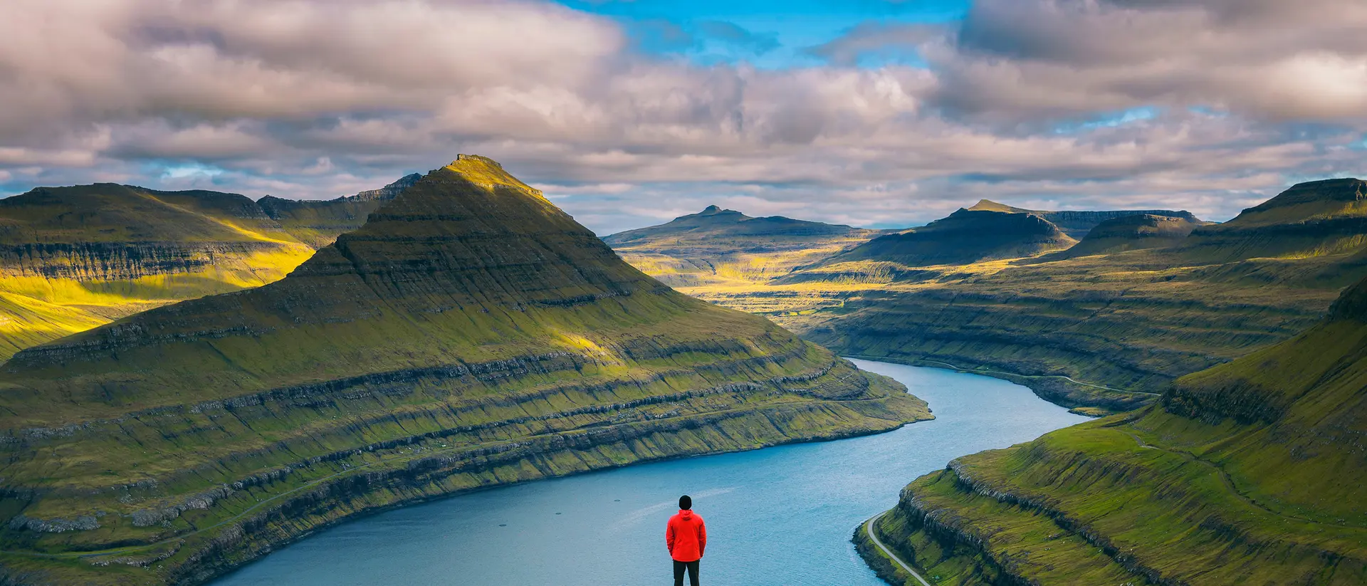 A person in a red jacket stands on a cliff overlooking a winding river and lush green mountains under a cloudy sky.