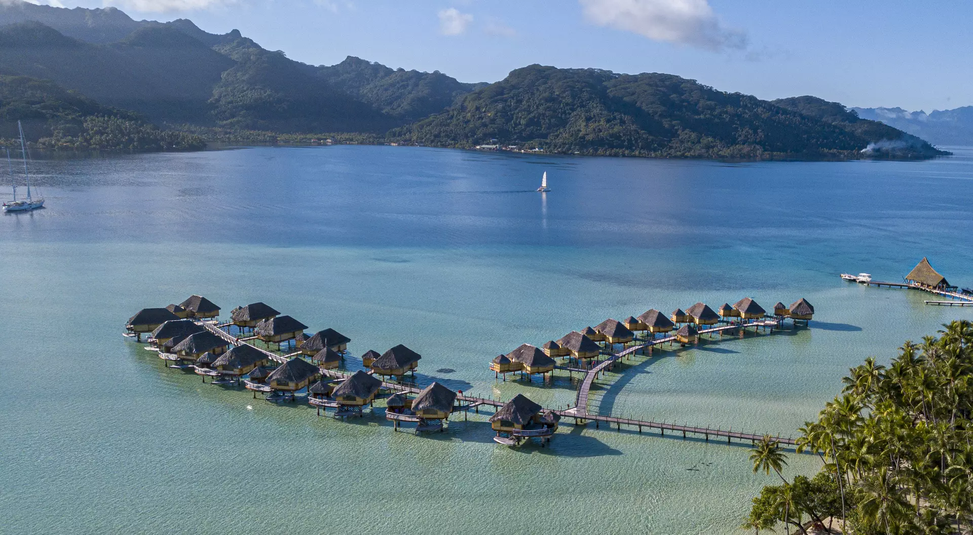 Overwater bungalows arranged in a curve, surrounded by clear turquoise water and lush green mountains in the background.