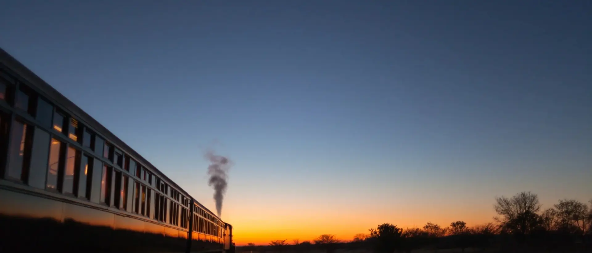 A steam train travels along tracks at sunset, with vibrant colors in the sky and a silhouette of trees in the distance.