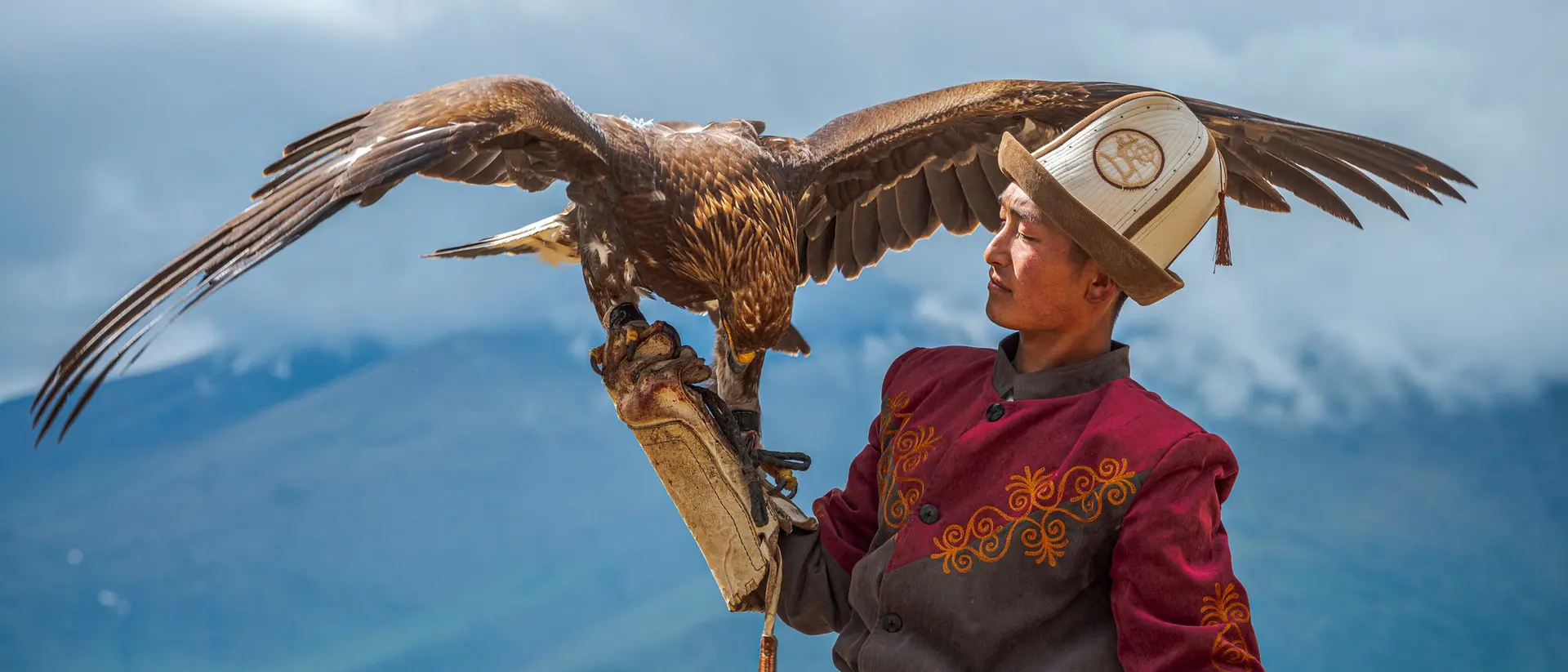 A man in traditional attire holds a large eagle on his arm against a mountainous backdrop.