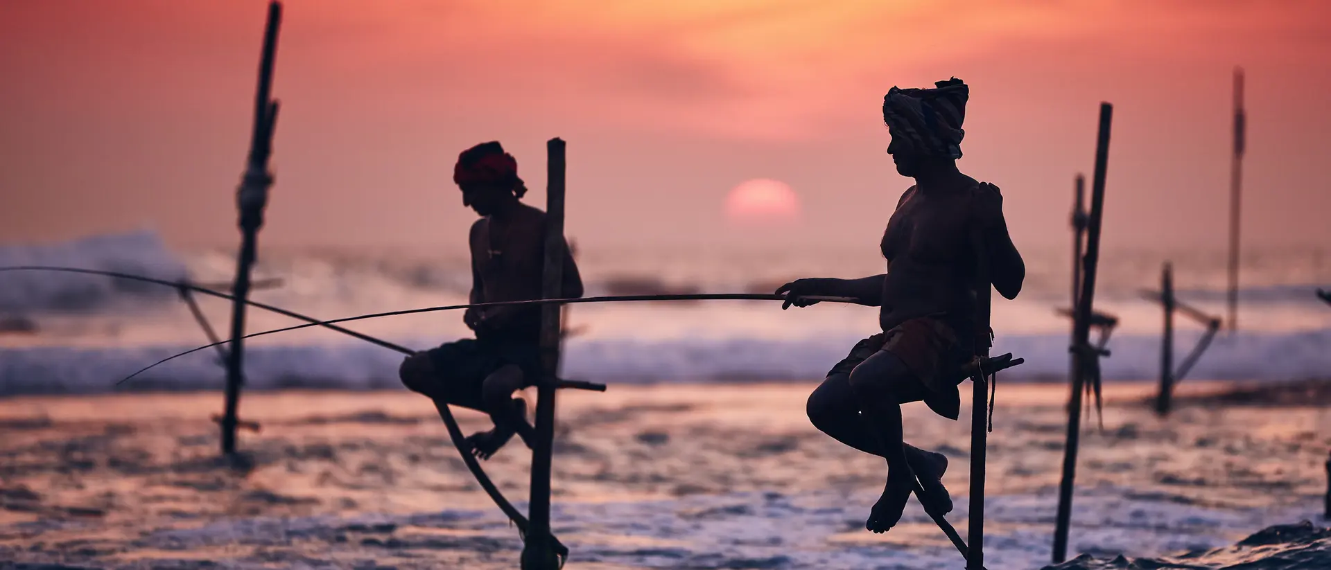 Silhouetted fishermen perched on poles in the ocean at sunset, casting lines into the water.