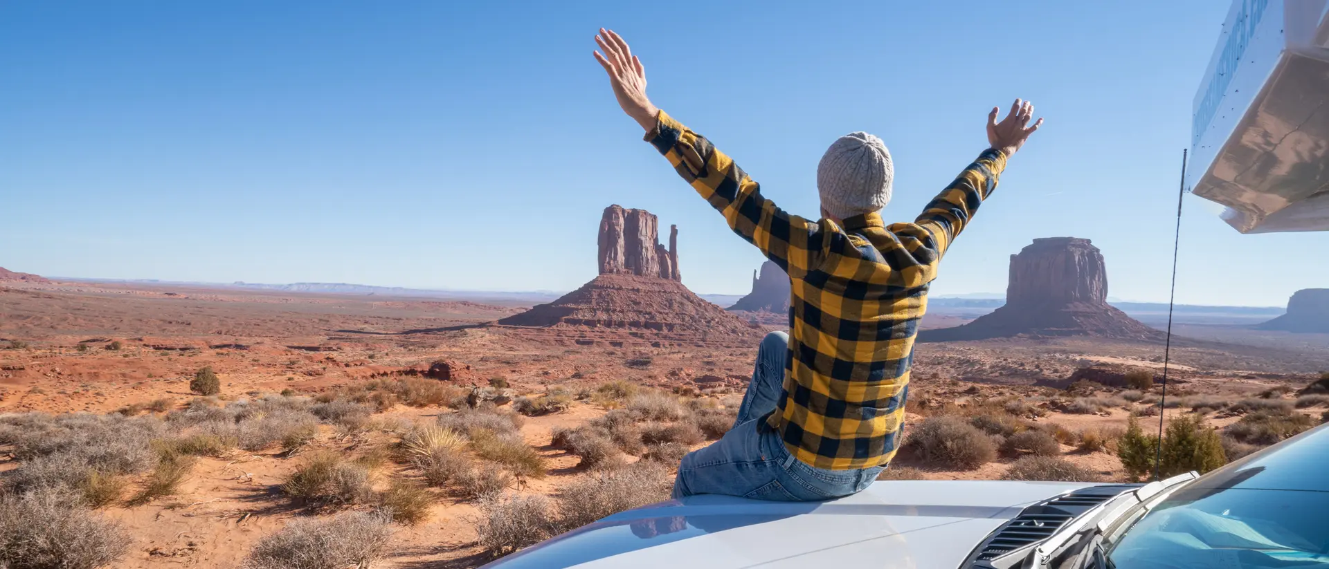 Person in a flannel shirt sits on a car roof, arms raised, overlooking Monument Valley's iconic buttes. Bright blue sky.