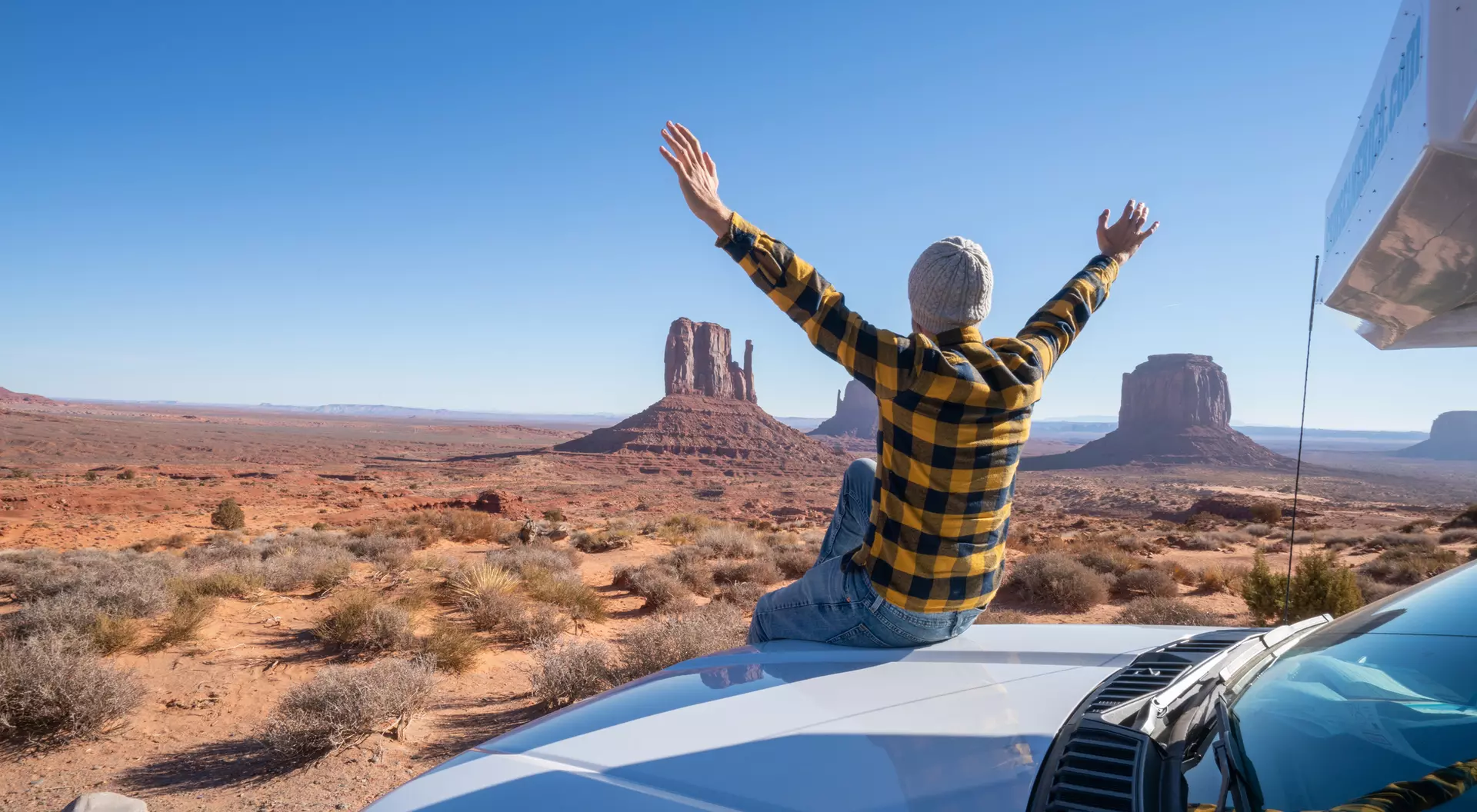 Person in a flannel shirt sits on a car roof, arms raised, overlooking Monument Valley's iconic buttes. Bright blue sky.