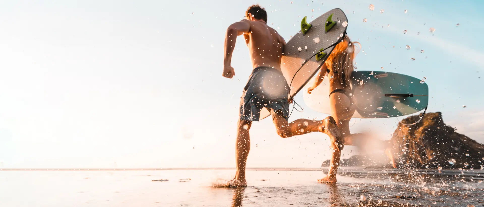 Two surfers run across a wet beach, carrying surfboards, as water splashes around them under a bright sky.