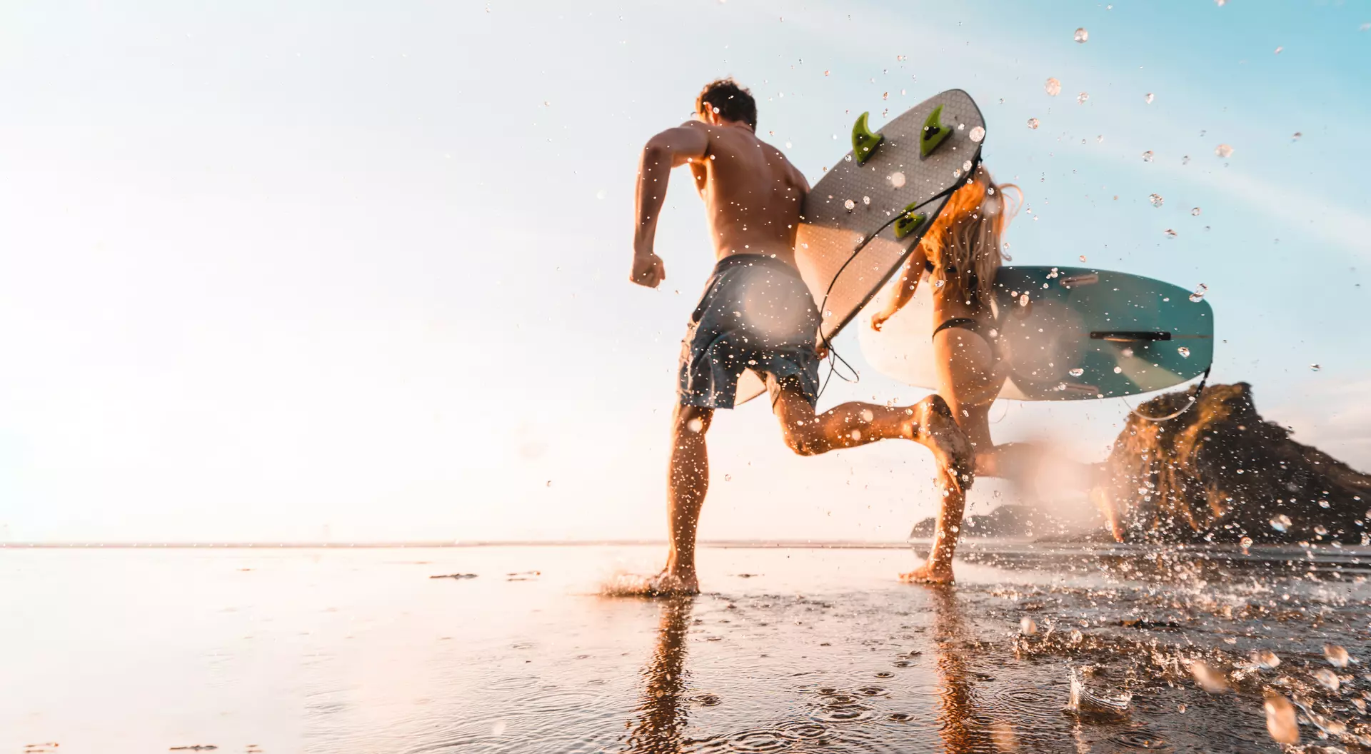 Two surfers run across a wet beach, carrying surfboards, as water splashes around them under a bright sky.