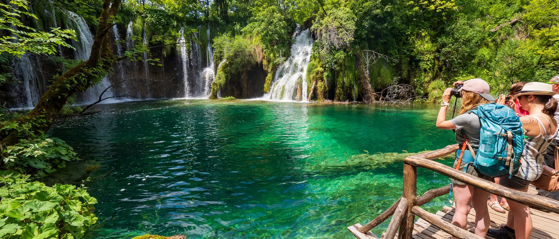 Two people stand on a wooden platform, taking photos of a turquoise lake and waterfalls surrounded by lush greenery.