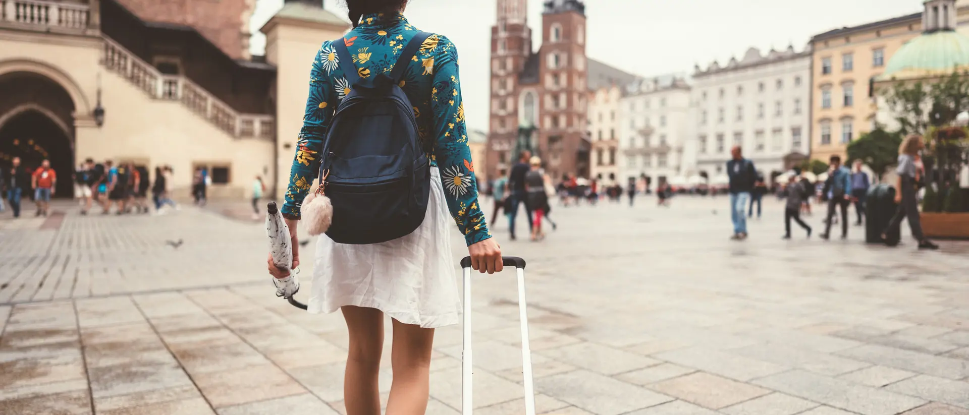 A person walks away, pulling a yellow suitcase through a bustling square with historic buildings in the background.