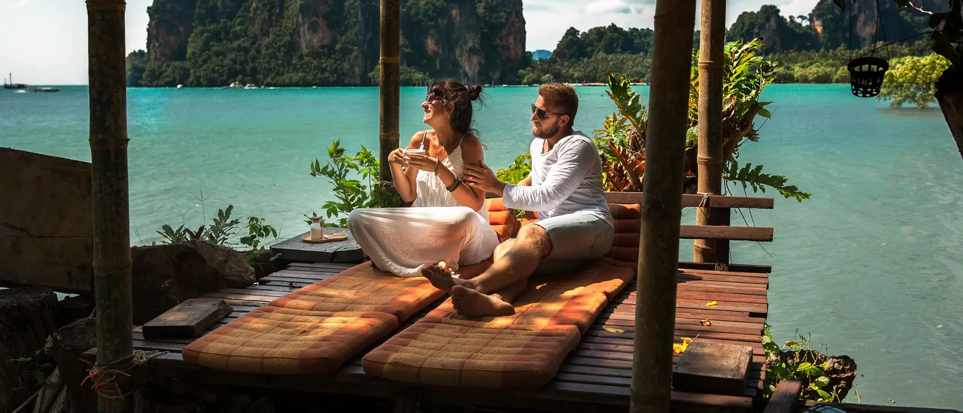 A couple sits on a wooden deck by the ocean, enjoying the view of distant mountains and lush greenery.