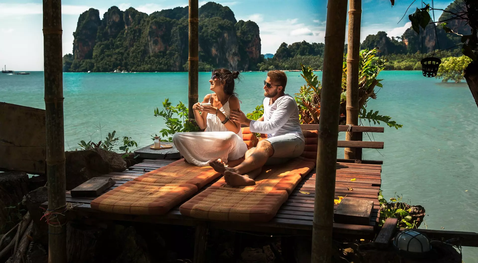 A couple sits on a wooden deck by the ocean, enjoying the view of distant mountains and lush greenery.