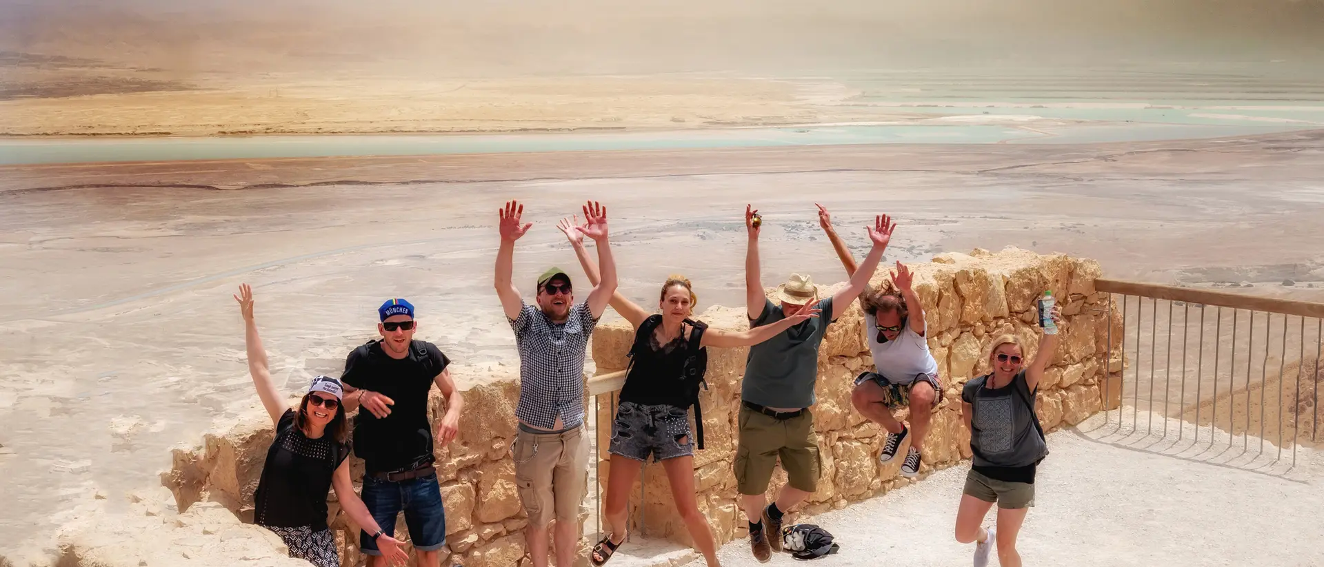 A group of seven people joyfully jumping in the air with a scenic view of the Dead Sea in the background.