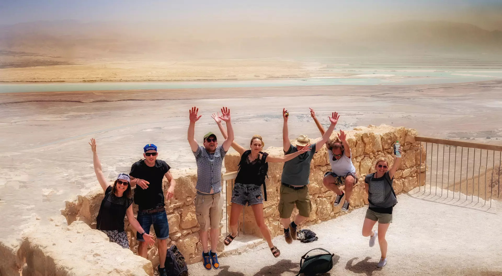 A group of seven people joyfully jumping in the air with a scenic view of the Dead Sea in the background.