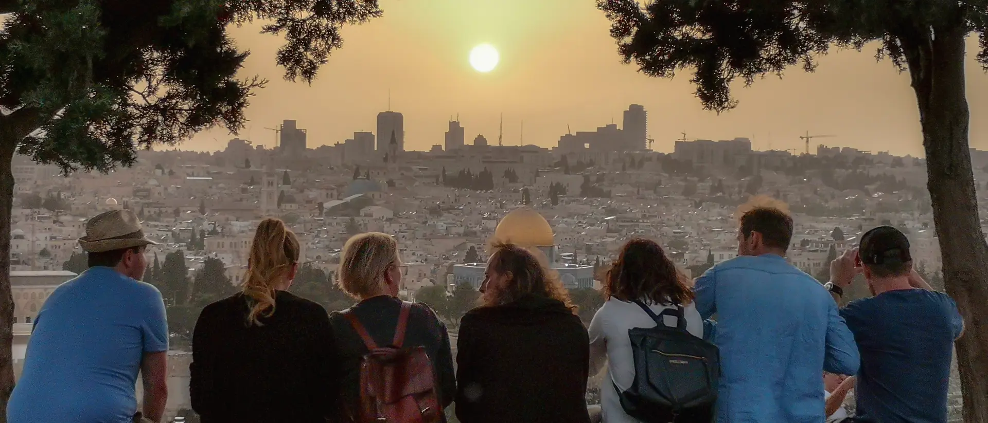 A group of people sit on a stone wall, watching the sunset over a city skyline. Trees frame the scene.