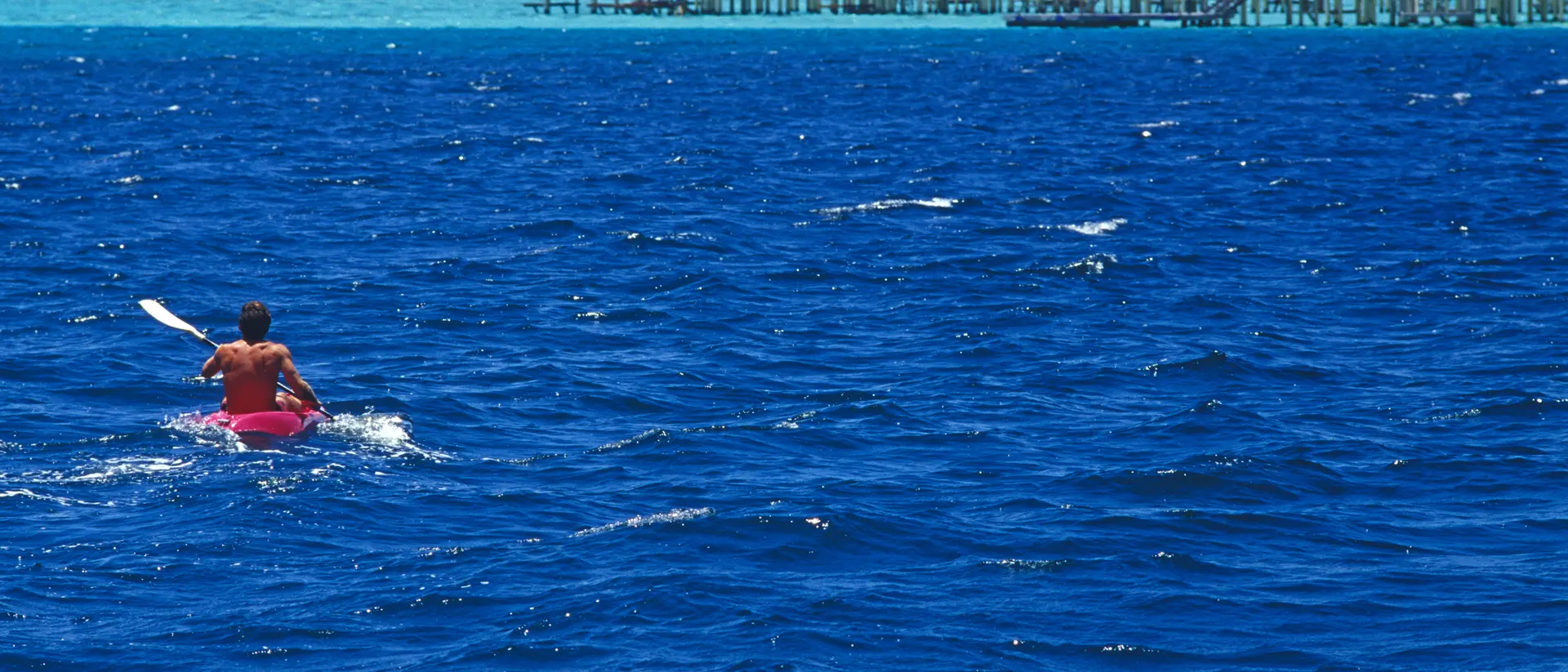 A person kayaks on bright blue water near overwater bungalows under a clear sky.
