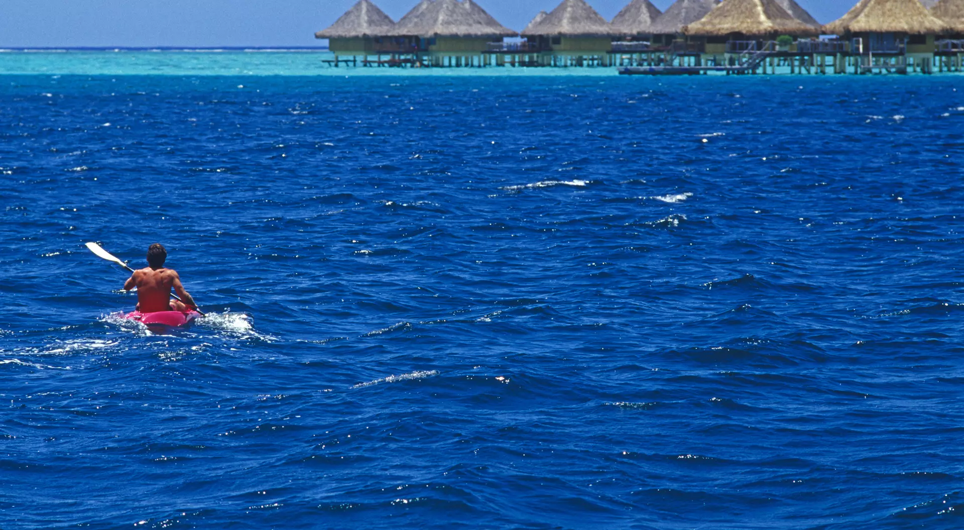 A person kayaks on bright blue water near overwater bungalows under a clear sky.