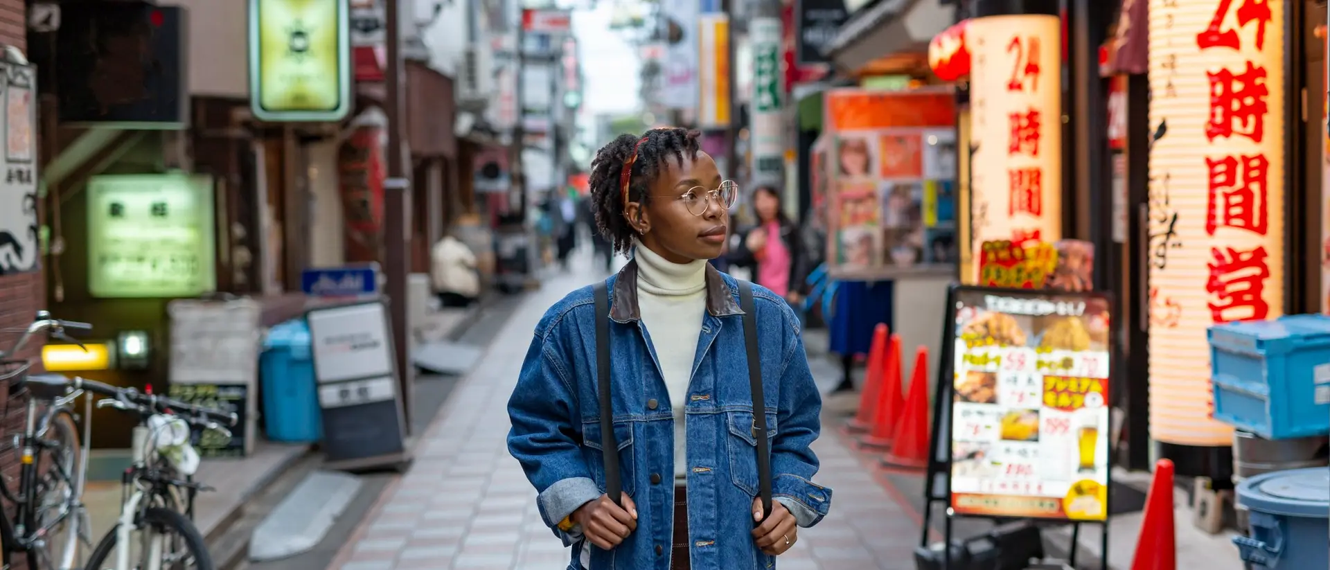 A person in a denim jacket stands in a vibrant urban alley filled with shops and neon signs.