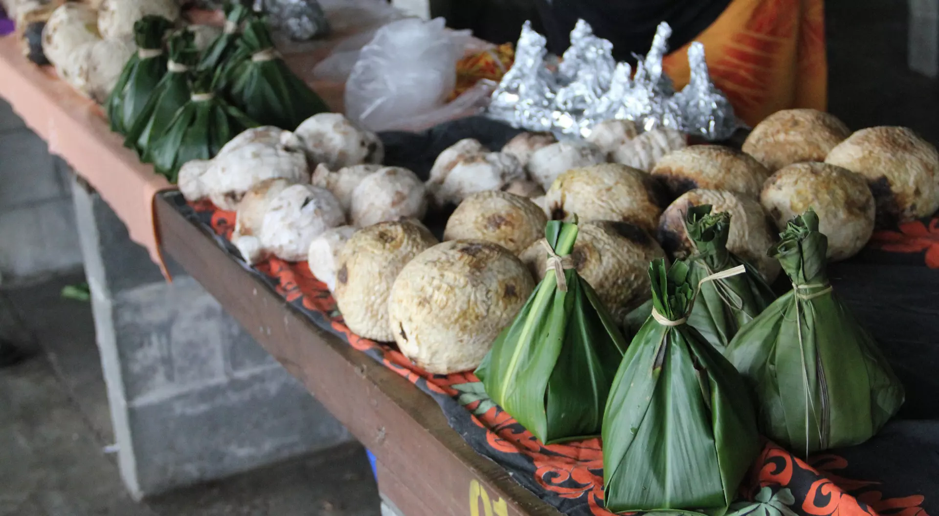 A market stall displays various food items, including round dumplings and bundled parcels wrapped in green leaves.