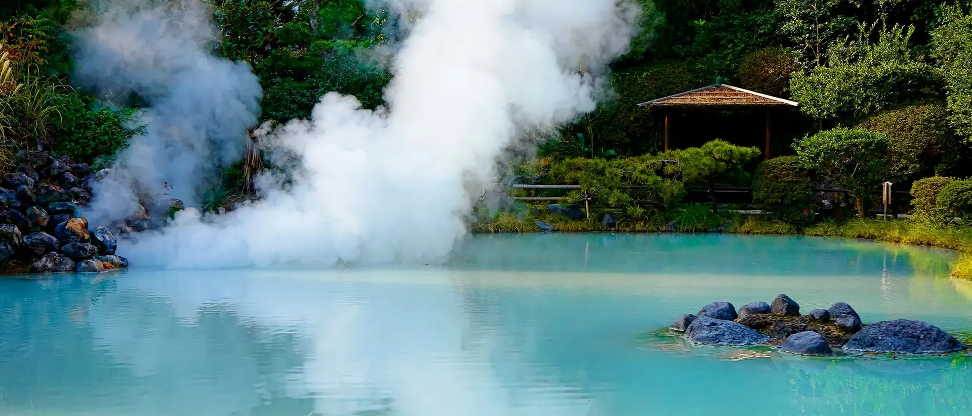 A serene hot spring with light blue water and steam rising, surrounded by lush greenery and a gazebo in the background.