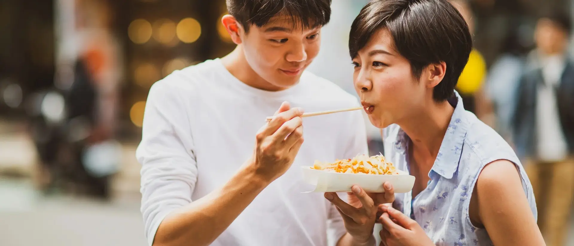 A young man feeding a young woman noodles on a bustling street, both smiling and enjoying the moment.