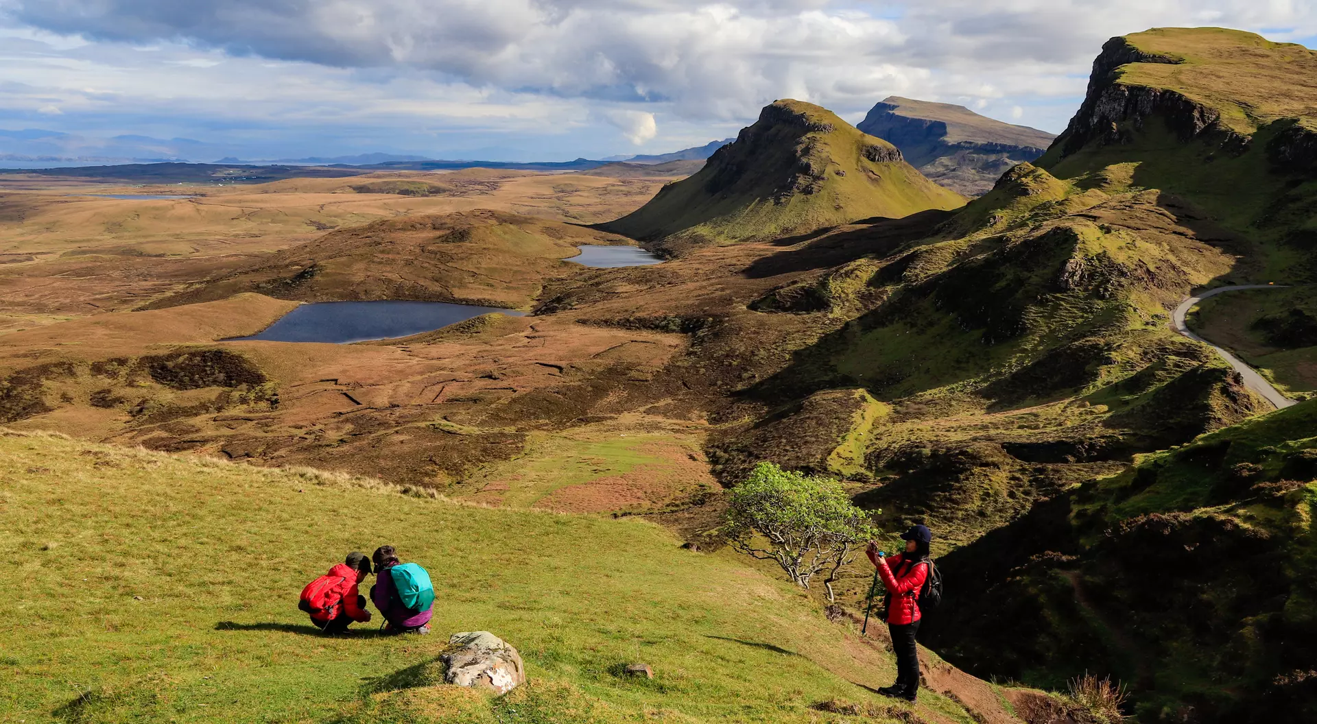 Two hikers explore a rocky hillside with a panoramic view of distant mountains and a tranquil landscape.