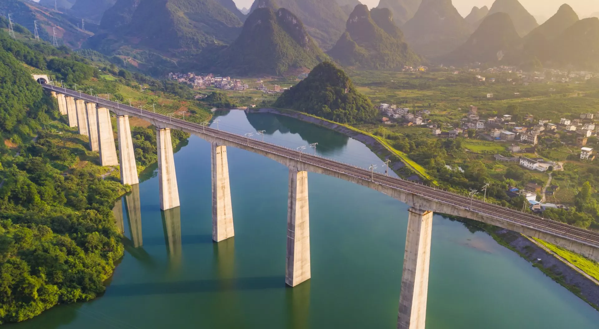 A railway bridge spans a river, surrounded by green hills and mountains under a soft sunrise glow.