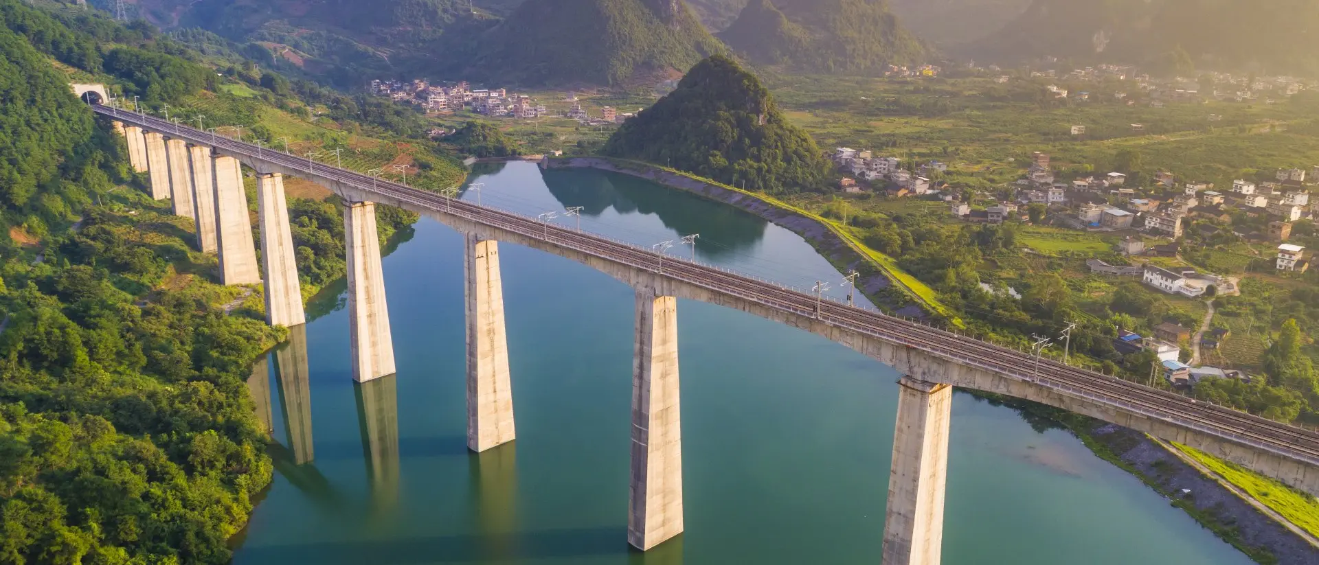 A railway bridge spans a river, surrounded by green hills and mountains under a soft sunrise glow.