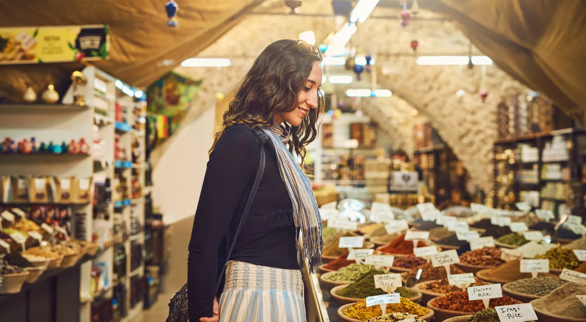 A woman stands in a market, admiring colorful spice displays in bowls, surrounded by shelves filled with various products.