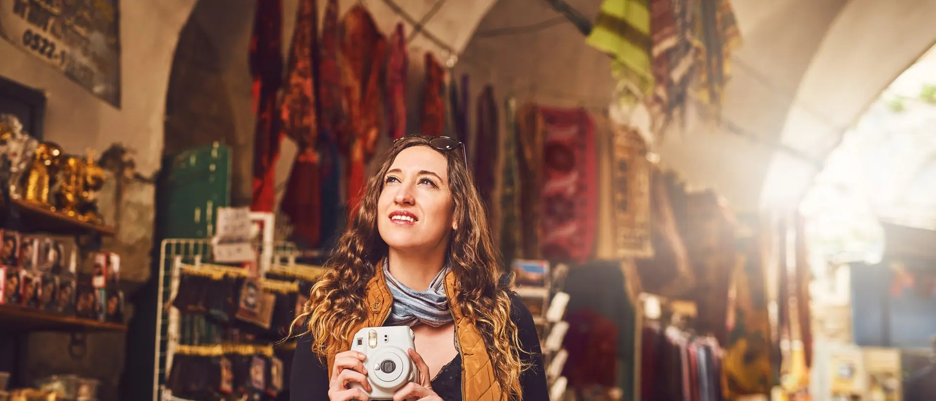 A woman with curly hair smiles while holding a camera, surrounded by colorful fabrics and crafts in a market.