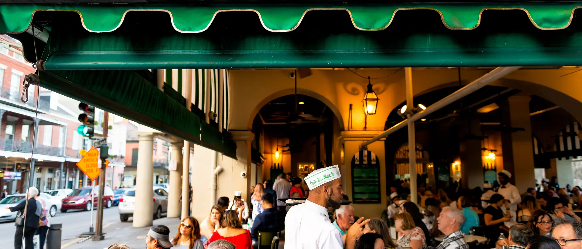 Outdoor scene at Cafe du Monde with diners at tables and a waiter in a white uniform. Green awning above with restaurant name.
