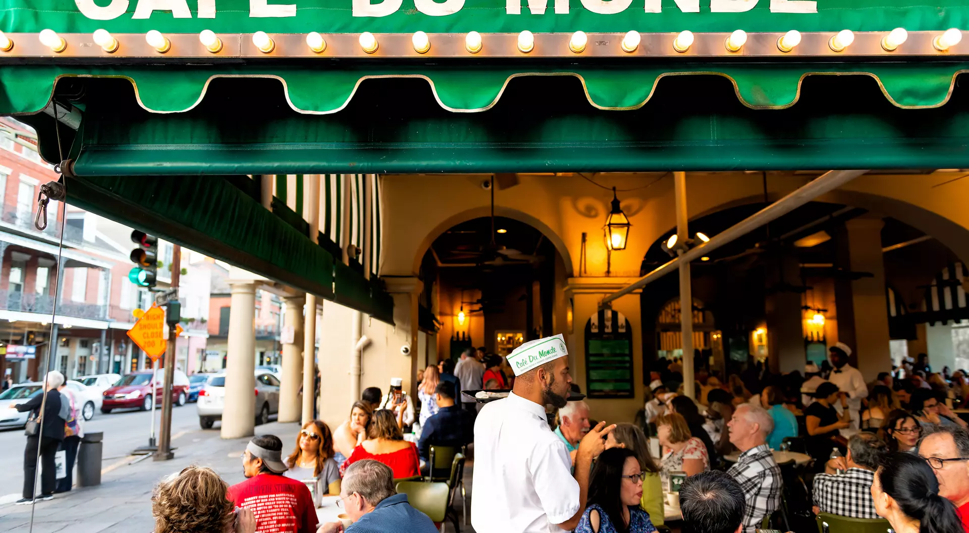 Outdoor scene at Cafe du Monde with diners at tables and a waiter in a white uniform. Green awning above with restaurant name.