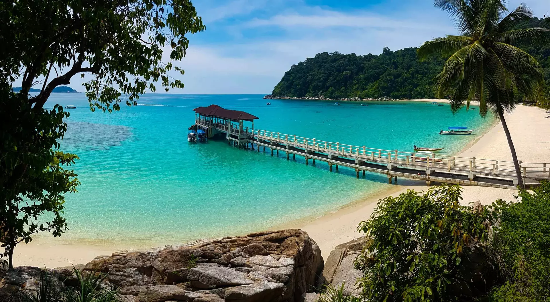 A scenic beach with a wooden pier extending into turquoise waters, surrounded by lush greenery and mountains in the background.