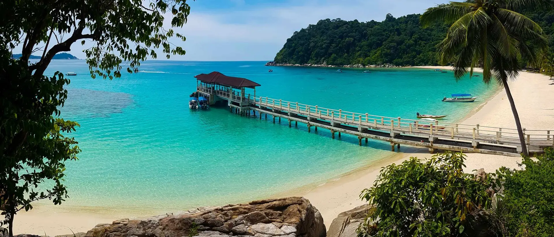 A scenic beach with a wooden pier extending into turquoise waters, surrounded by lush greenery and mountains in the background.