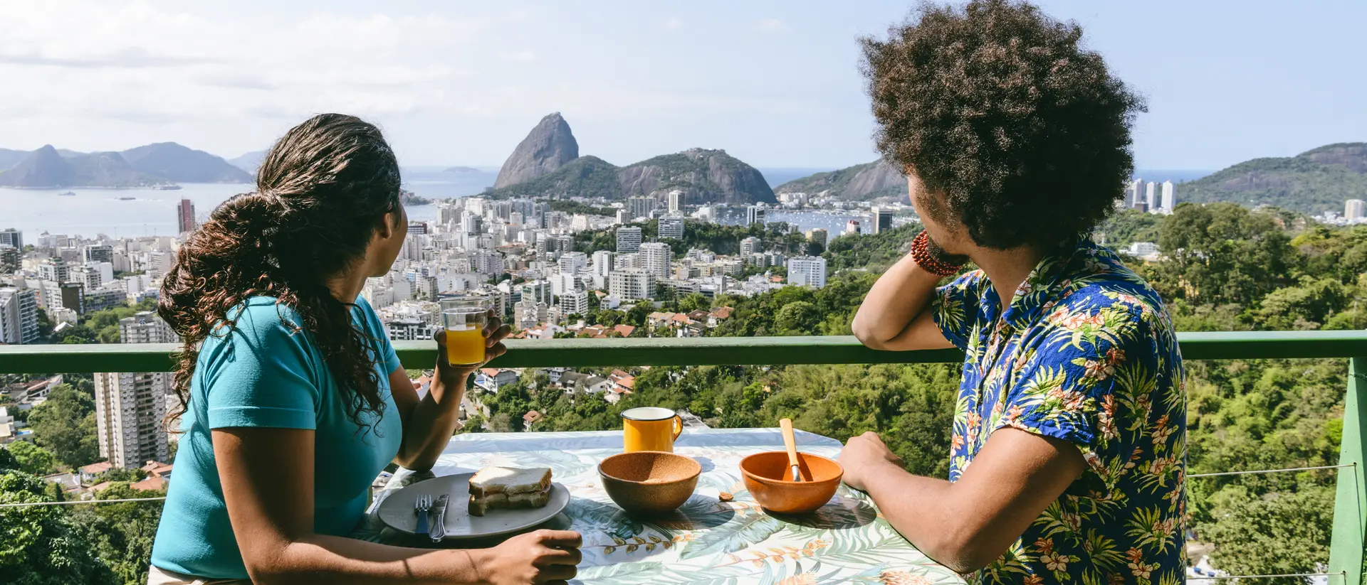 Two people sit at a table with bowls and drinks, overlooking a city and mountainous landscape.