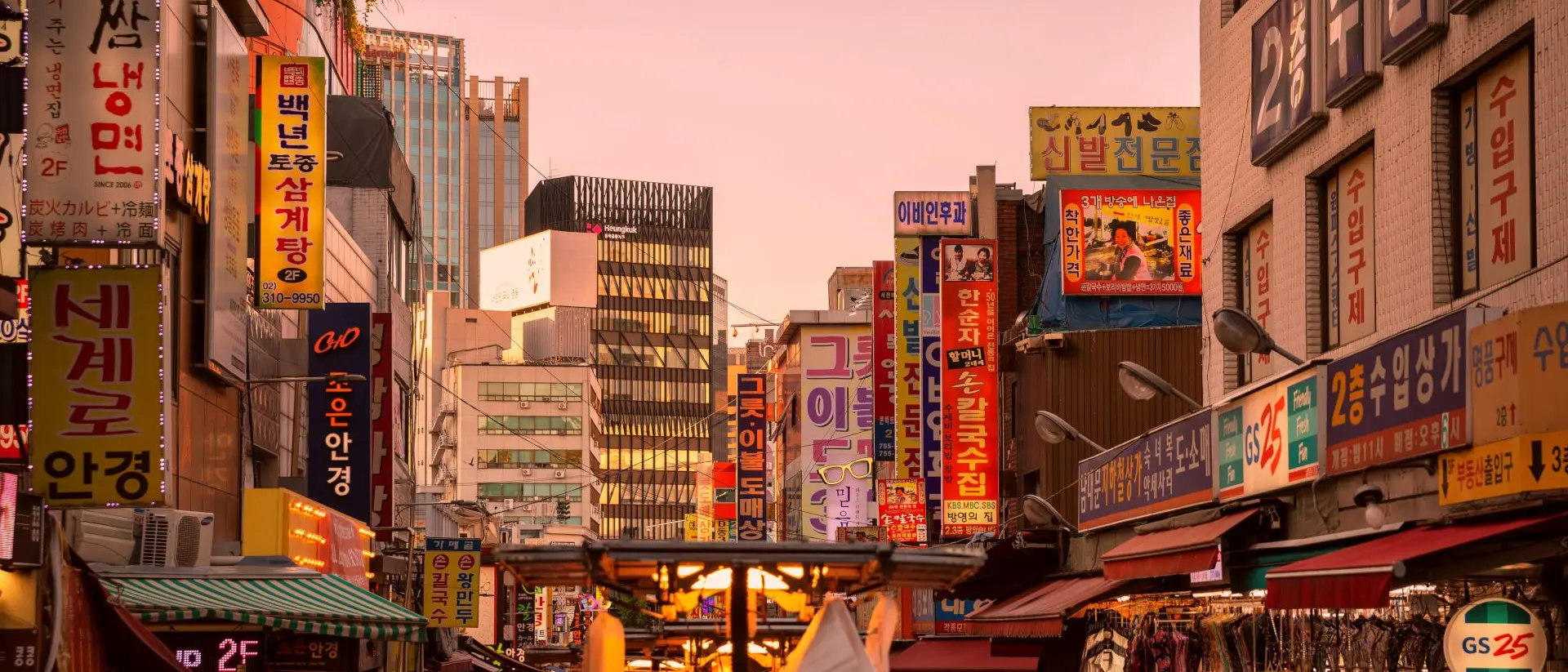 A bustling street market at dusk, filled with colorful signs and people shopping among stalls.