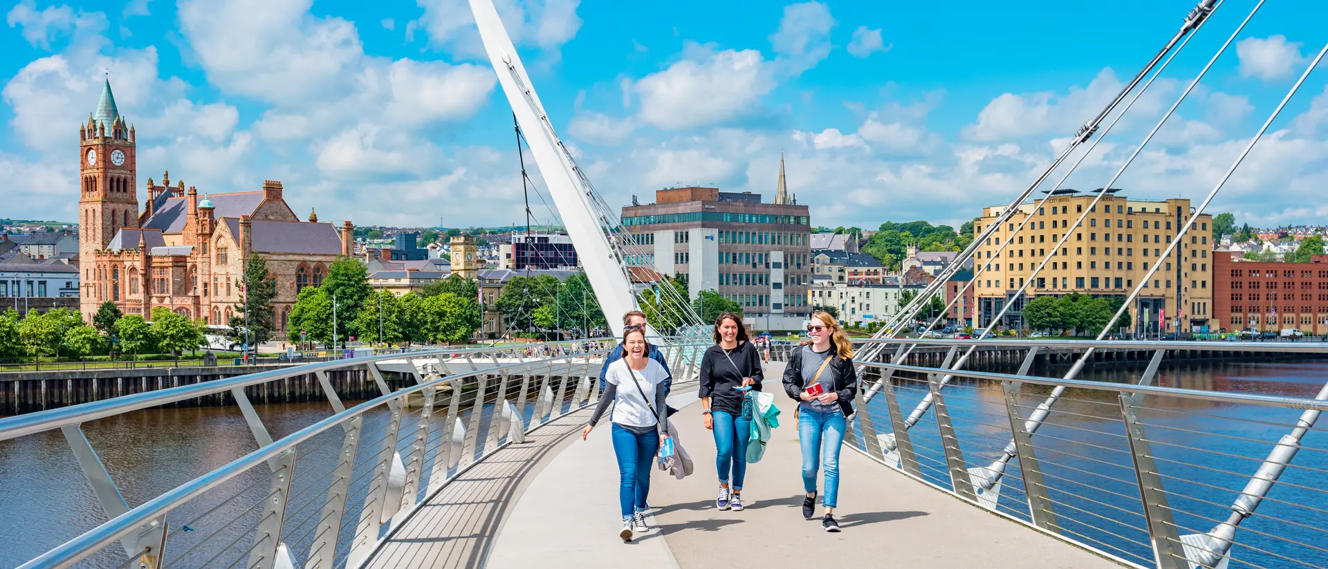 Four people walk across a bridge with a city skyline and blue sky in the background.