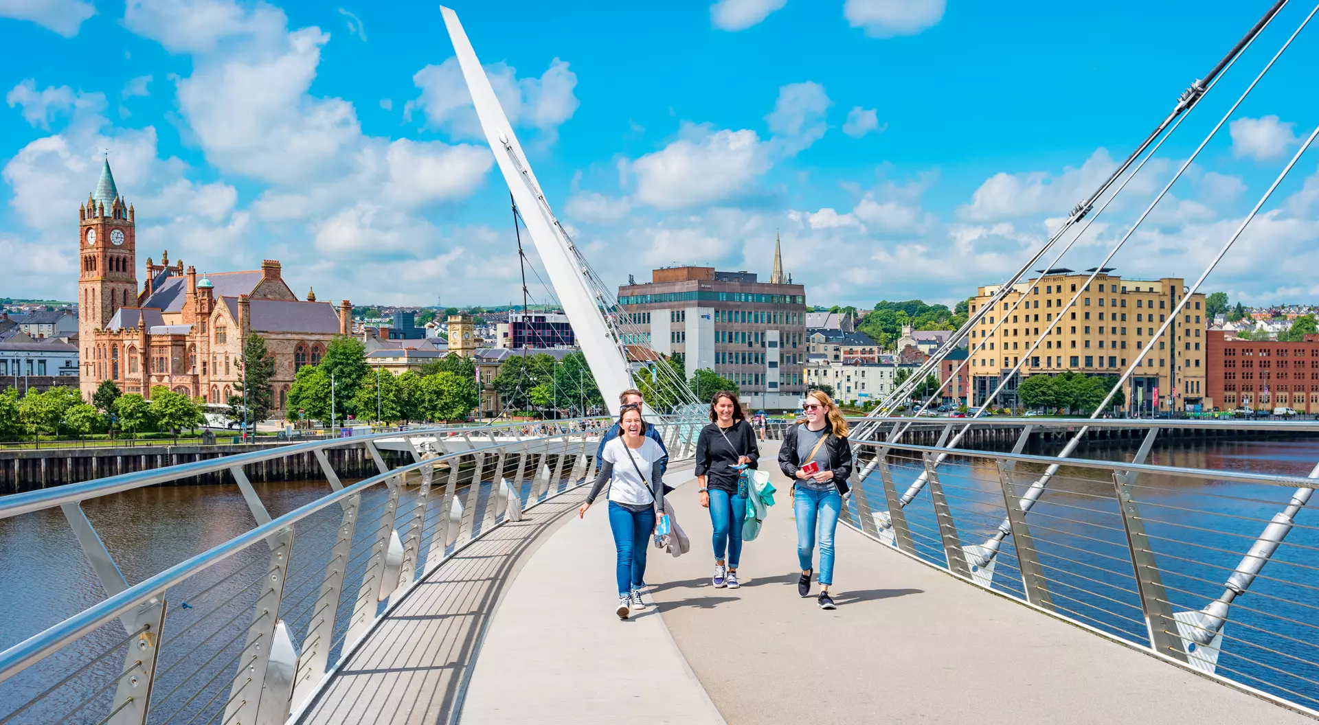 Four people walk across a bridge with a city skyline and blue sky in the background.