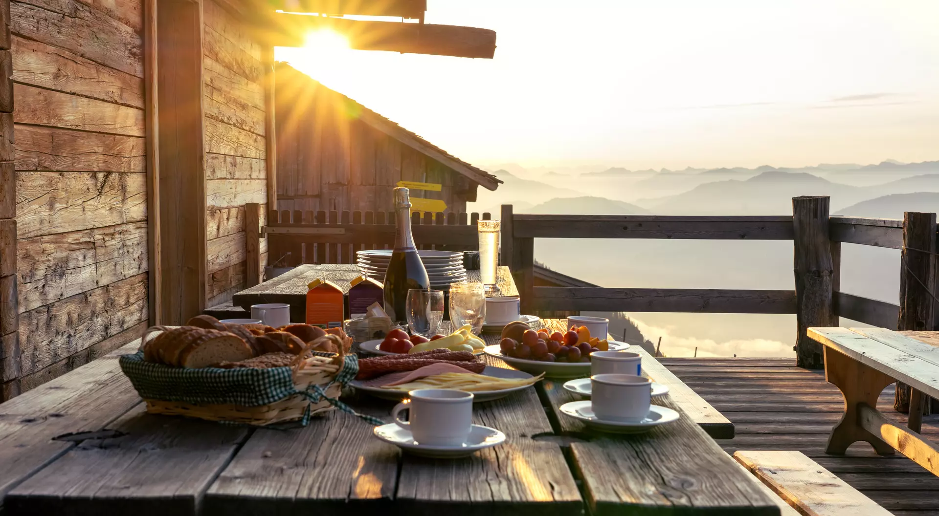 Breakfast spread on a wooden table with coffee, pastries, and fruit, illuminated by sunrise over misty mountains.