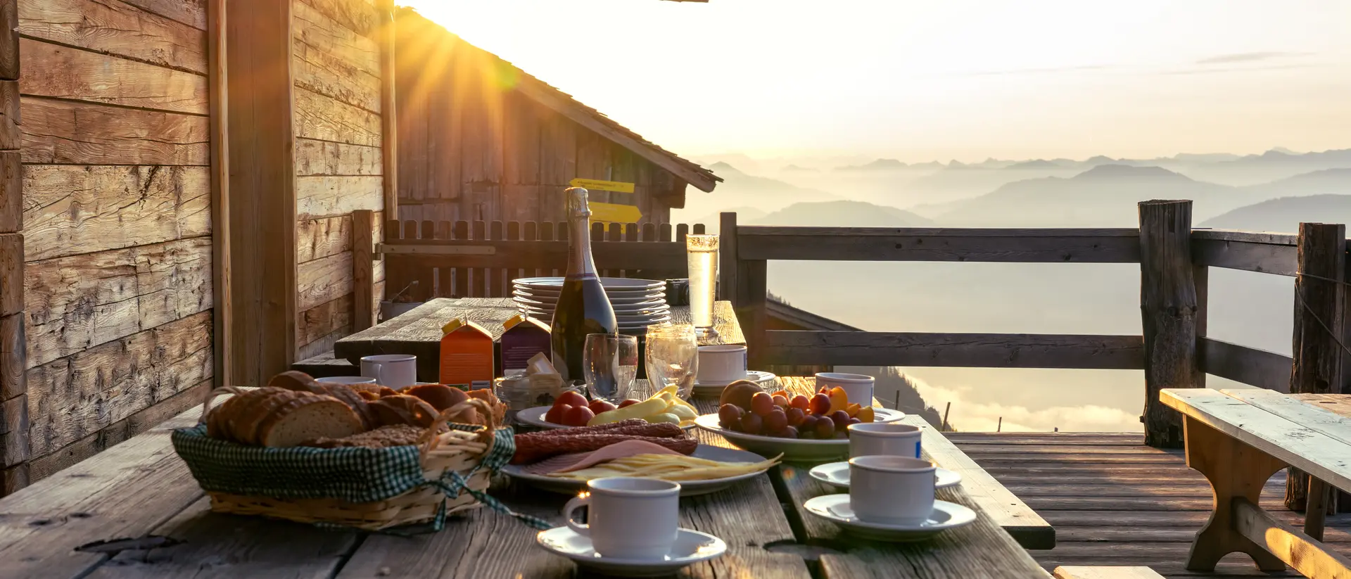 Breakfast spread on a wooden table with coffee, pastries, and fruit, illuminated by sunrise over misty mountains.