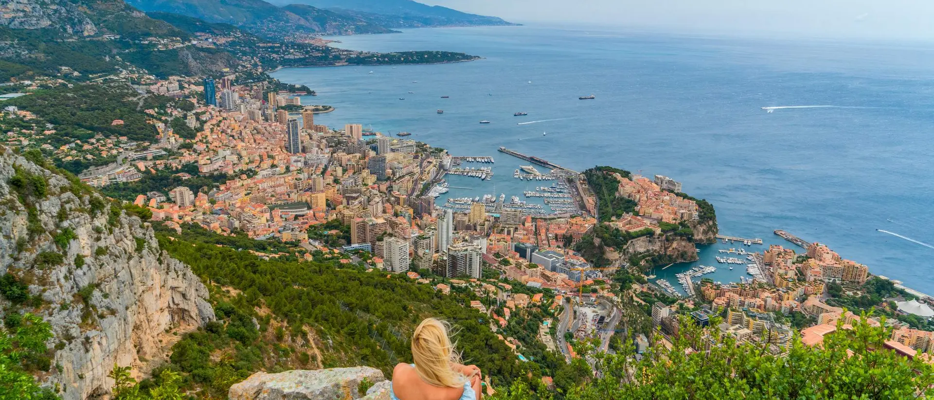A woman sits on rocks overlooking a coastal city, with buildings and boats nestled along the shoreline and hills in the background.